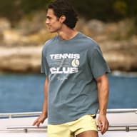 Man wearing a blue Tennis Club graphic T-shirt and yellow shorts outdoors by the sea.