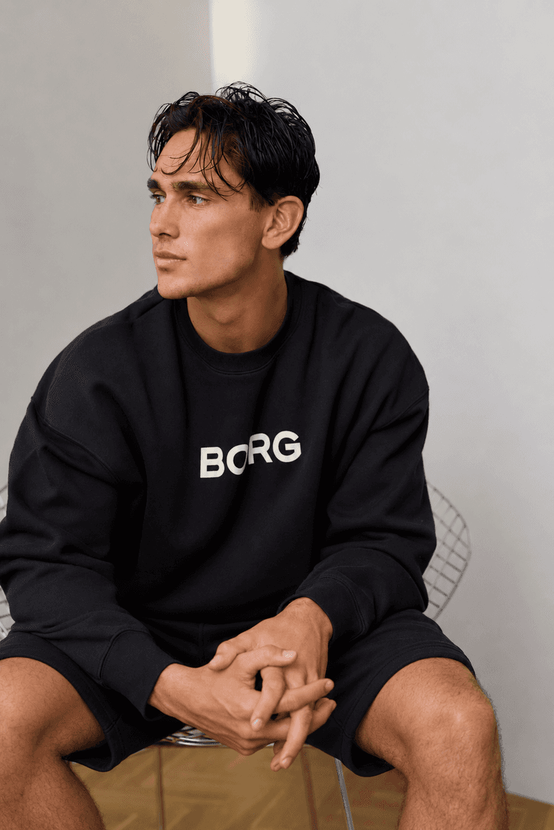Male model seated in a black Björn Borg sweatshirt and matching black shorts