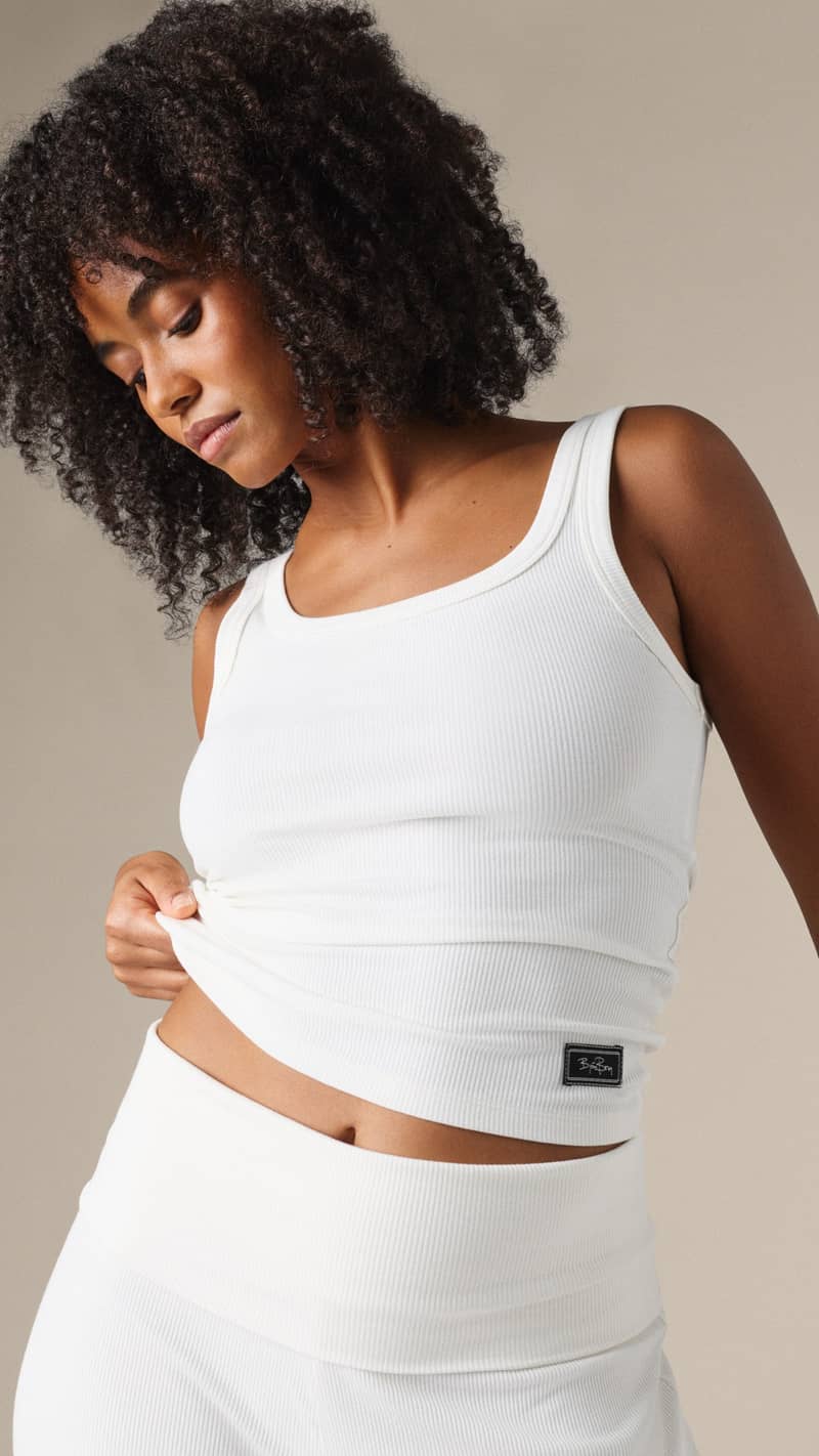 A woman with curly hair wearing a white ribbed tank top and bottom, looking down, set against a neutral background.