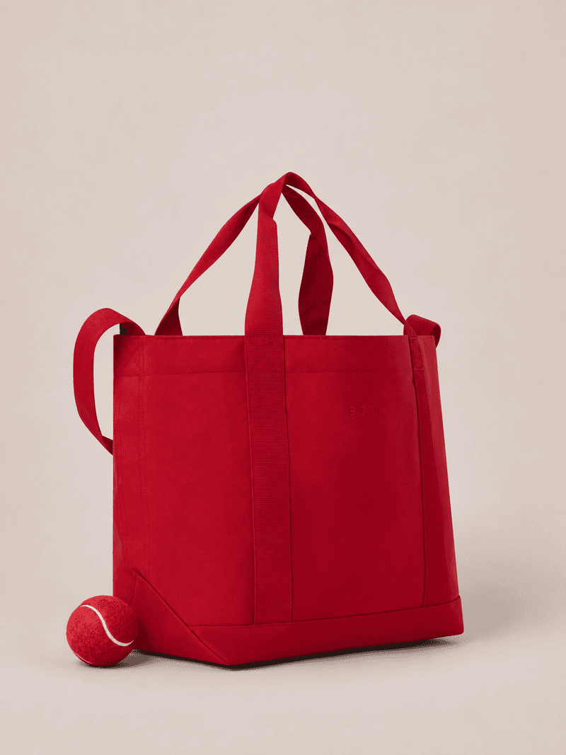 Red tote bag with matching straps and a textured red ball beside it against a neutral background.