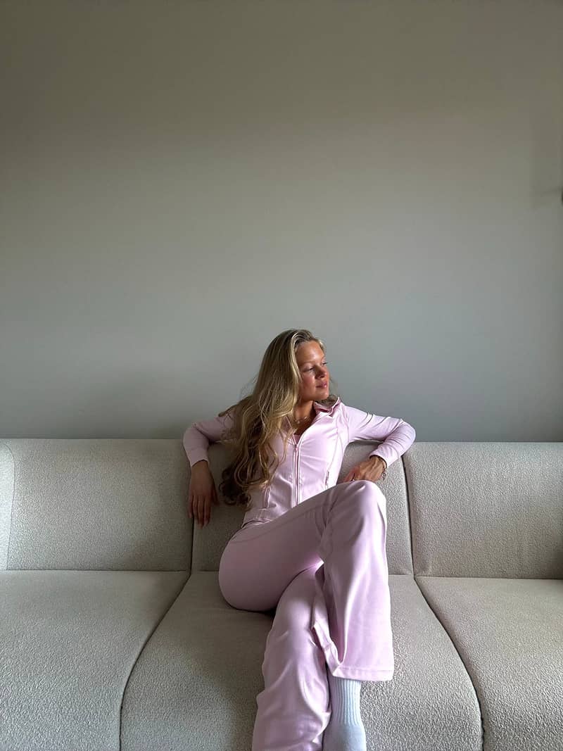 A woman wearing a pink zip midlayer jacket and matching tights, is sitting on a light-colored sofa .