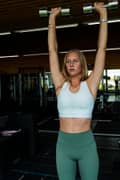 Woman doing dumbbell shoulder presses at the gym, wearing a white sports bra and green tights from Björn Borg