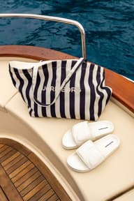 Striped Björn Borg tote bag and white slides on a boat seat, with a view of blue water in the background.