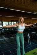 Woman doing lateral raises with dumbbells at the gym, wearing white sports bra and green gym leggings from Björn Borg