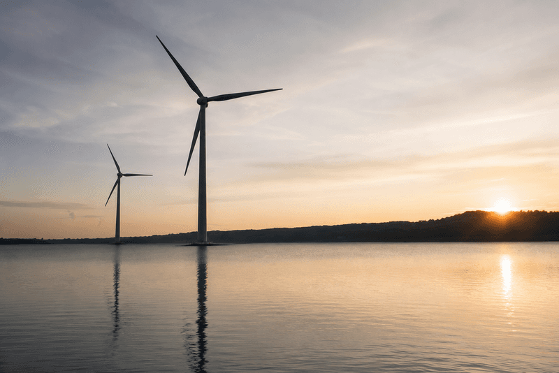 Two wind turbines stand in calm water during a serene sunset, with silhouetted hills in the background.