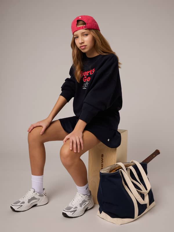 Teen seated on a wooden box wearing a red cap, black outfit, and sneakers. A navy tote with a tennis racket rests beside them.