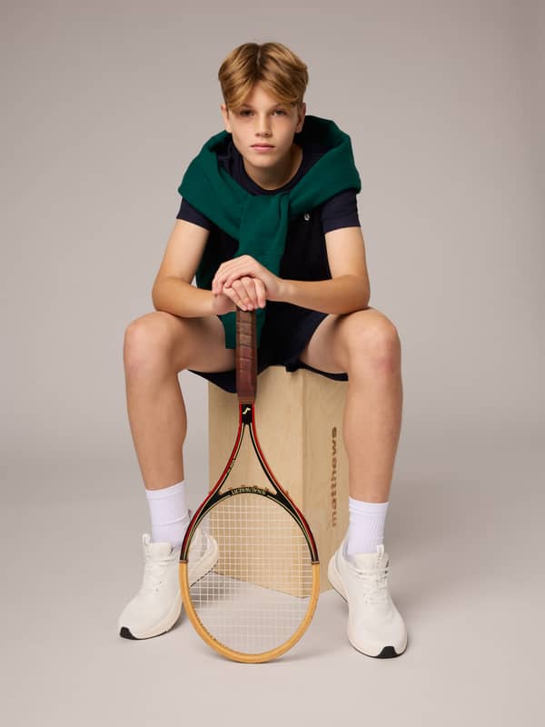 Young person sits on a box with a tennis racket, wearing a green sweater over a navy outfit and white sneakers, looking at the camera.