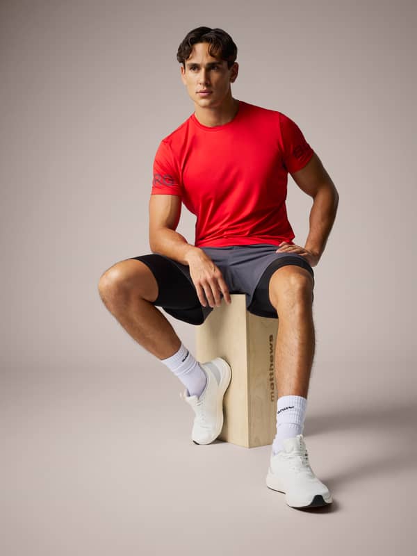 Man in a red T-shirt, gray shorts, and white sneakers sits on a wooden block in a studio setting.