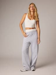 Woman with long blonde hair in a white crop top, gray sweatpants, and sneakers, posing against a neutral background.