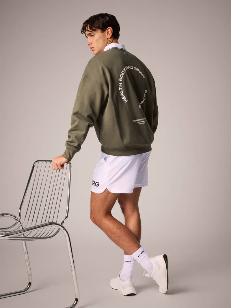 Man in olive sweatshirt and white shorts leans on a metal chair, showing sweatshirt's back print, against a plain background.