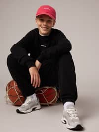 Smiling boy in a red cap and black outfit, sitting on basketballs, wearing gray sneakers against a neutral background.