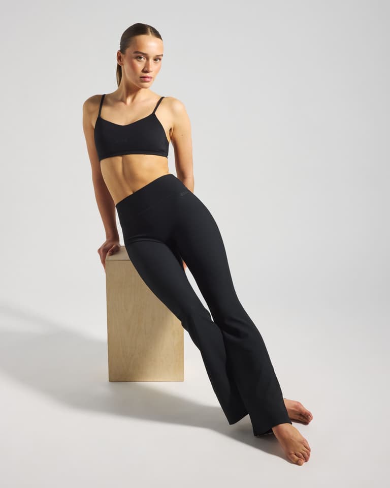 A woman in a black sports outfit leans on a wooden block in a minimalist setting, posing with a confident expression.
