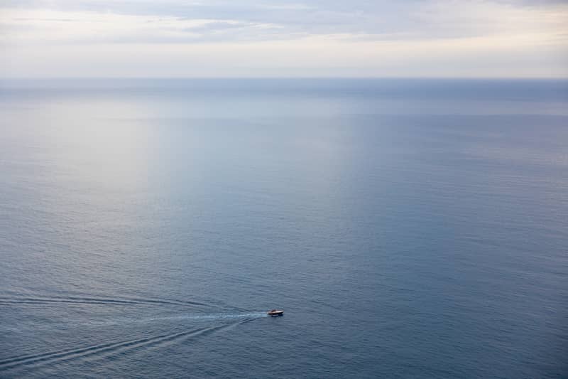 A small boat leaves a wake as it glides across a vast, calm ocean under a cloudy sky.