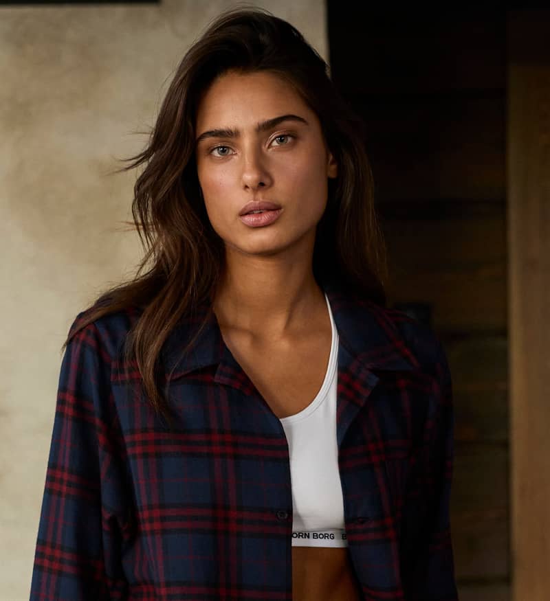A woman with long brown hair wearing a plaid shirt over a white sports bra stands indoors, looking directly at the camera.