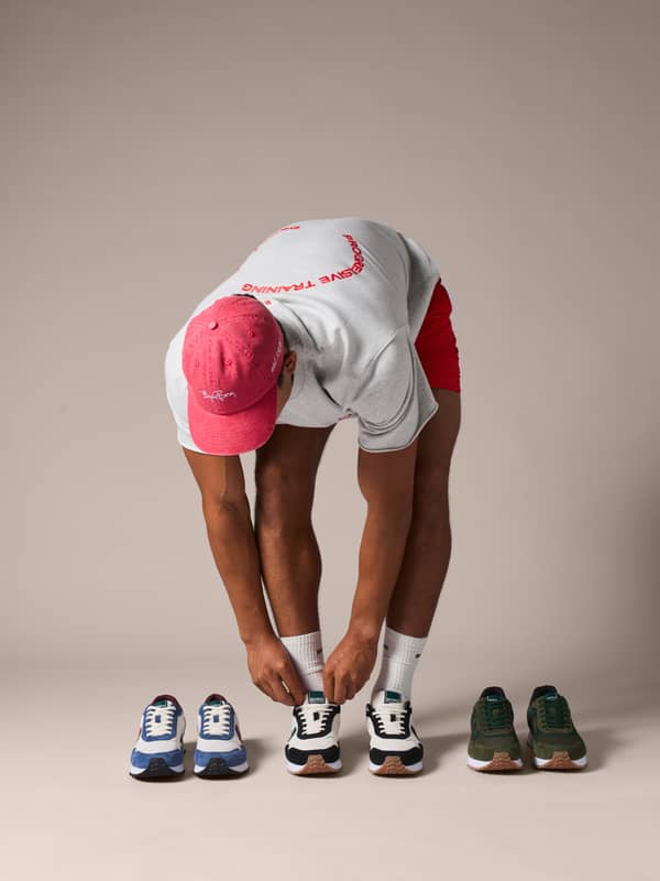 Person in a pink cap and gray shirt bends to tie sneakers, with additional pairs placed beside them on a neutral background.