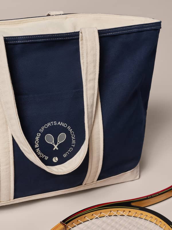 Cream and navy sports tote with "Sports and Racquet Club" logo, next to a vintage wooden tennis racquet on a beige background.