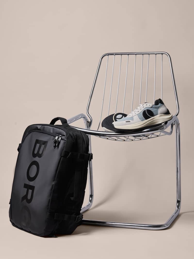A black backpack with "BORG" lettering on a metal chair, alongside white and gray sneakers, set against a plain background.