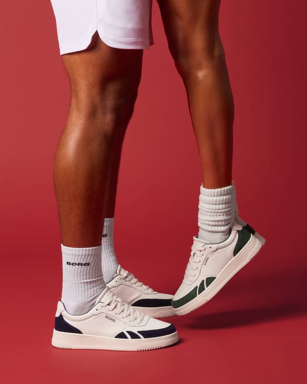 Two people wearing white sneakers with navy and green accents stand closely on a red background, one person on tiptoes.