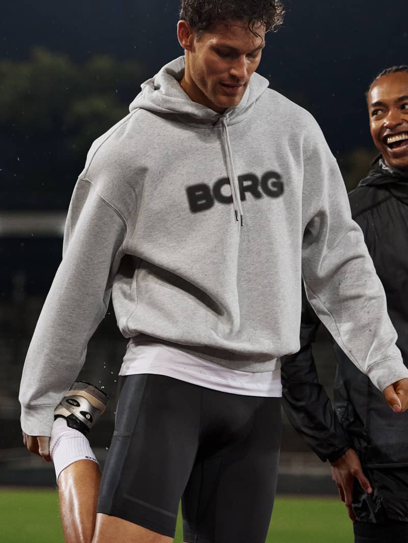 Man in a gray "BORG" hoodie and black shorts stands outdoors, smiling with friends, holding a soccer ball.