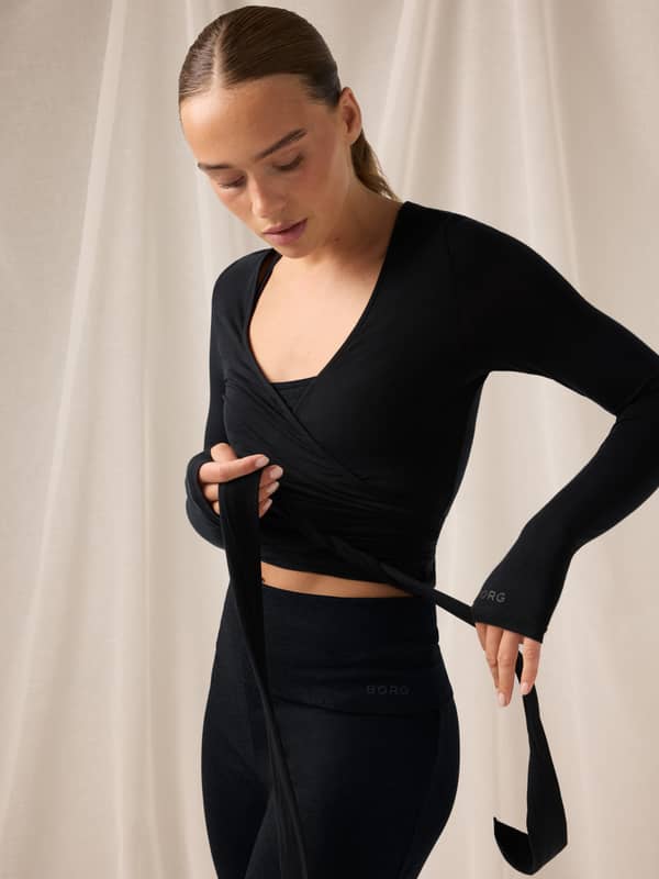 Woman in a black wrap top and leggings adjusts her clothing, standing against a light fabric backdrop with her hair tied back.