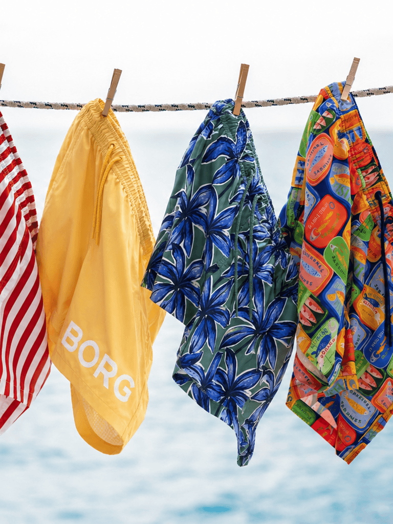 Four colorful swim trunks hang on a line, featuring red stripes, yellow, blue floral, and a vibrant pattern.