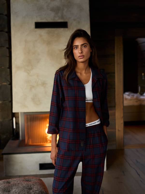 Woman wearing a navy and red checked pyjama set by a fireplace.