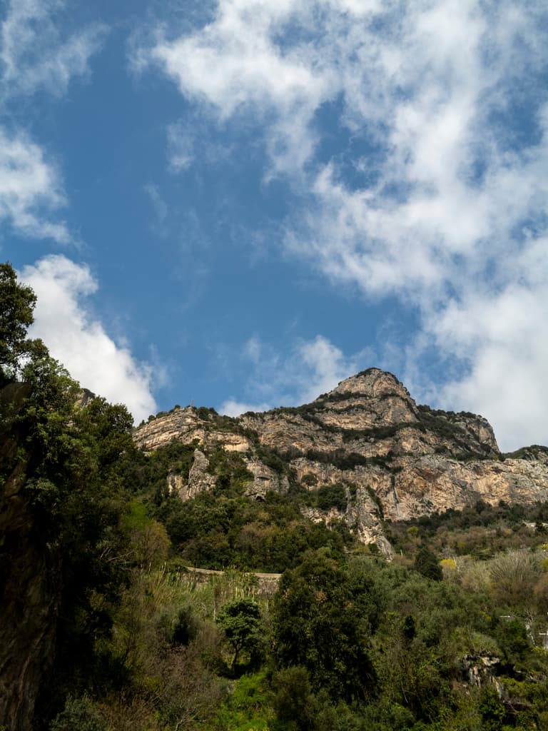 Mountain landscape with rocky peaks and lush greenery under a bright blue sky scattered with white clouds.