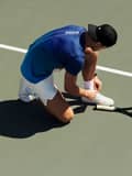 Tennis player in a blue shirt and white shorts kneels to tie his shoe on a court, with a racket lying nearby.