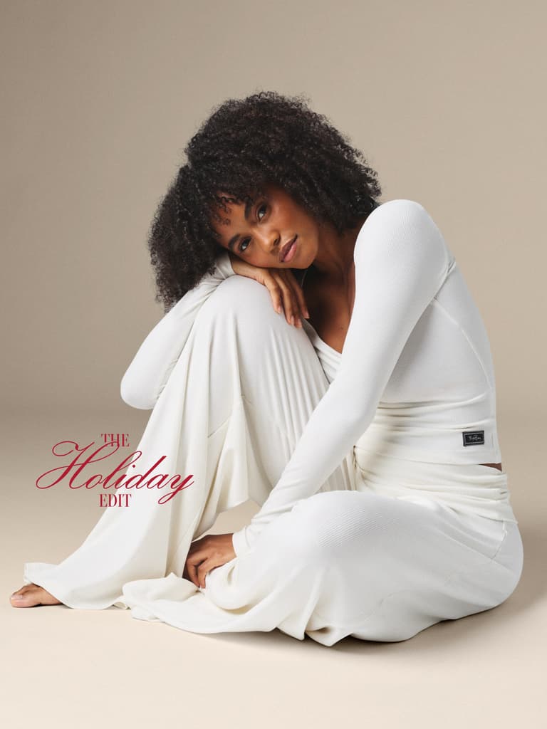 A woman with curly hair sits on the floor, wearing a white long-sleeve top and pants, resting her head on her knee, with "The Holiday Edit" text beside her.