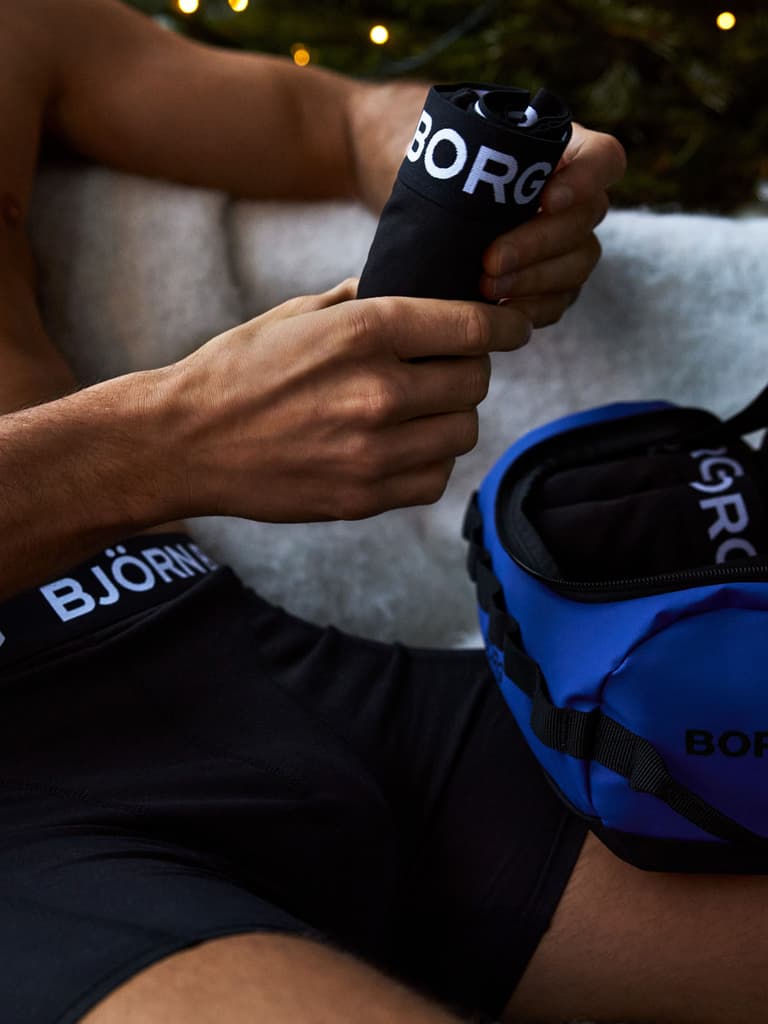 Man sitting indoors holding black Björn Borg underwear beside a blue bag.