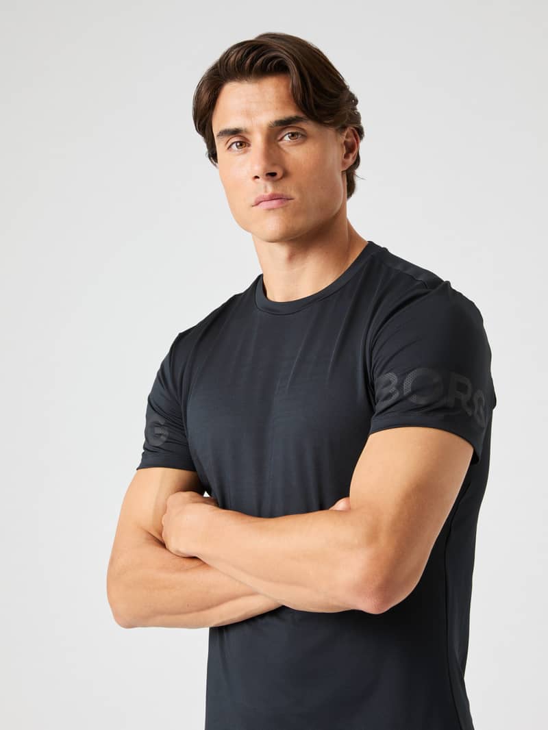Man in black athletic shirt with crossed arms, neutral expression, against a plain background.