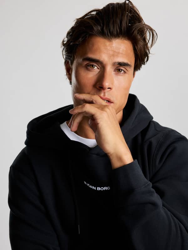 A man with tousled hair in a black hoodie with a thoughtful expression, resting his chin on his hand against a neutral background.