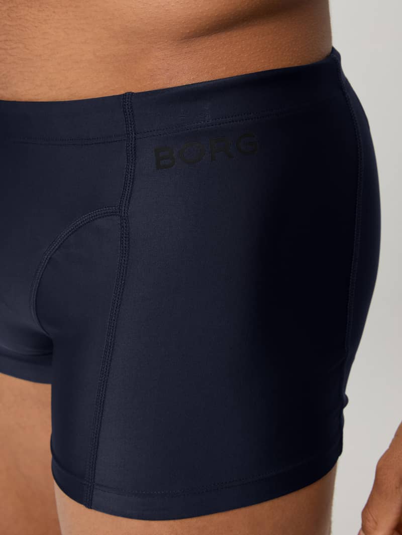 Close-up of a person wearing navy blue fitted swim trunks with "BORG" printed on the side.