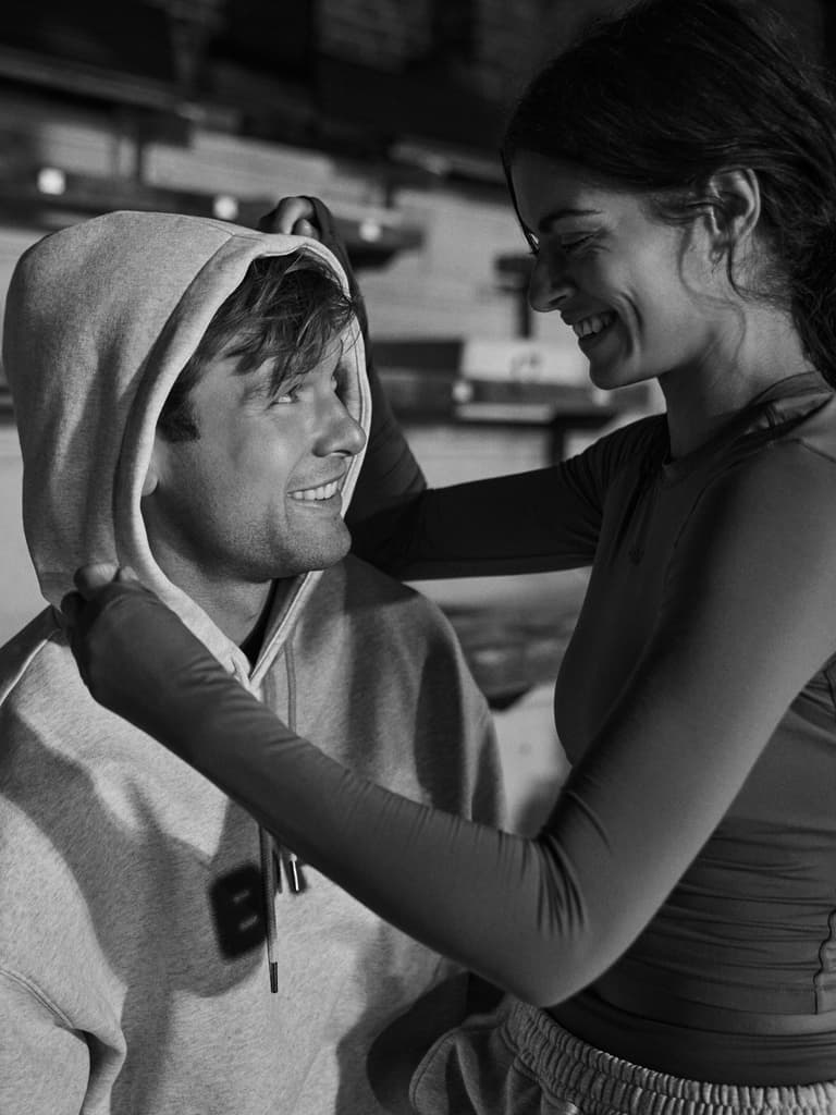 Woman playfully adjusts man's hoodie as they smile at each other, sitting in a cozy, casual setting. Black and white image.