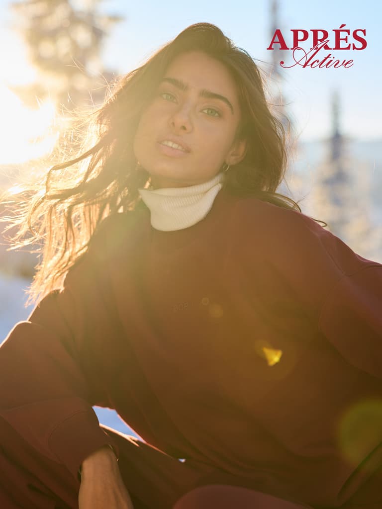 Woman in sunlight with flowing hair and a red outfit, wearing a turtleneck. Snowy background with logo text "APPRÉS Active" in the corner.