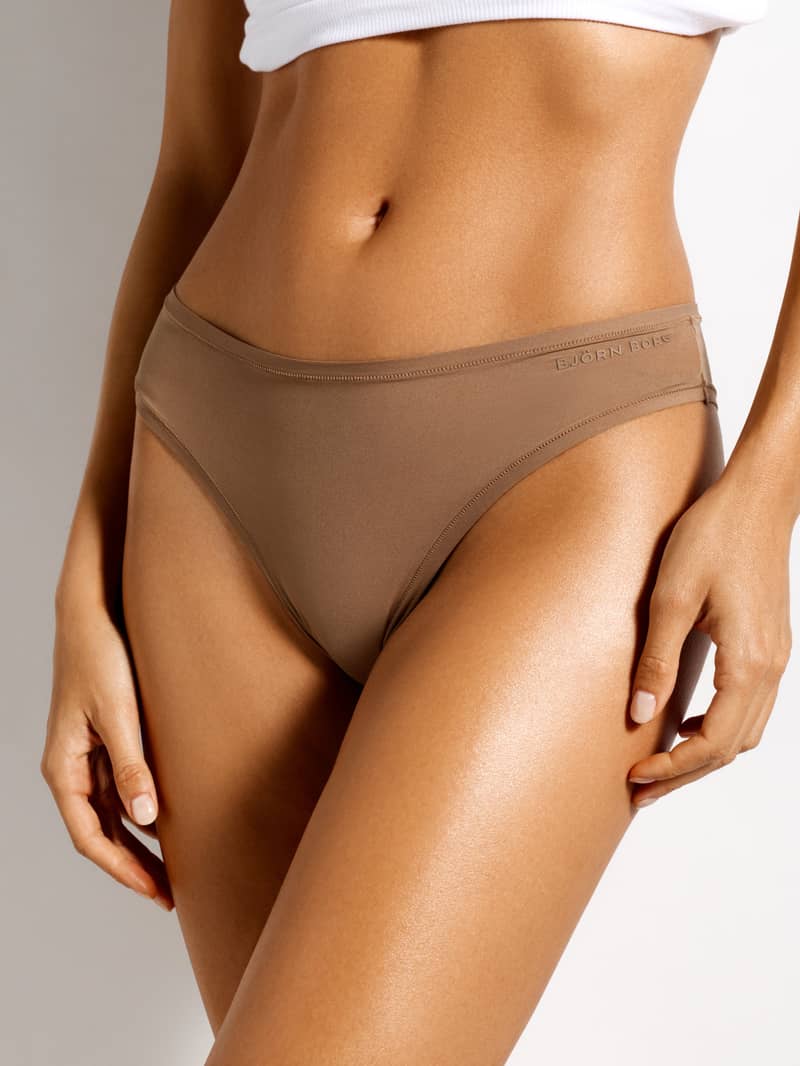 A woman in a white top wears brown Björn Borg underwear, with her hand resting on her thigh.