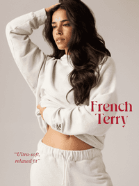 A person in a white French terry lounge set with relaxed fit, standing confidently, one hand on head, styled with long dark hair.