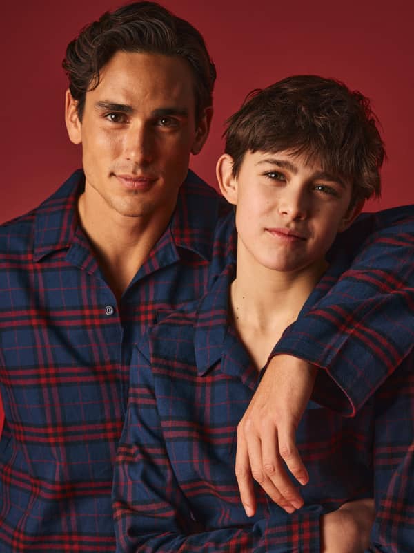Two people in matching blue and red plaid shirts, smiling against a red background, with one person's arm resting on the other's shoulder.