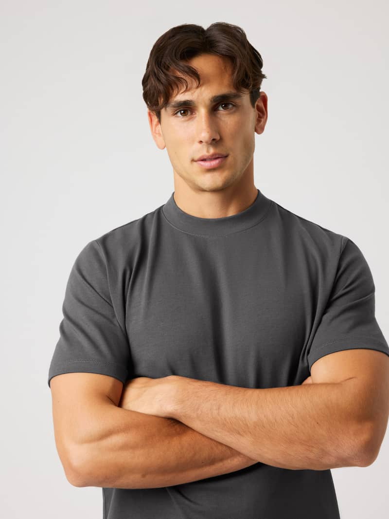 Man with dark wavy hair, wearing a gray t-shirt, standing with arms crossed against a light gray background.