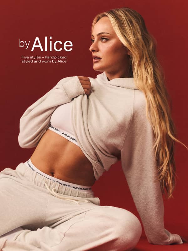 A woman in beige loungewear sits against a red backdrop, lifting her hoodie to reveal a white sports bra with Björn Borg branding.
