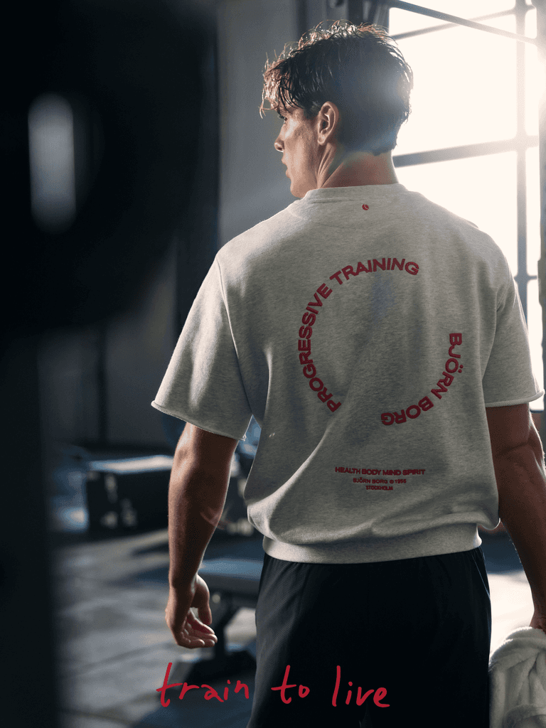A man stands in a gym wearing a gray shirt with red text and black shorts. The phrase "train to live" is written at the bottom in red.