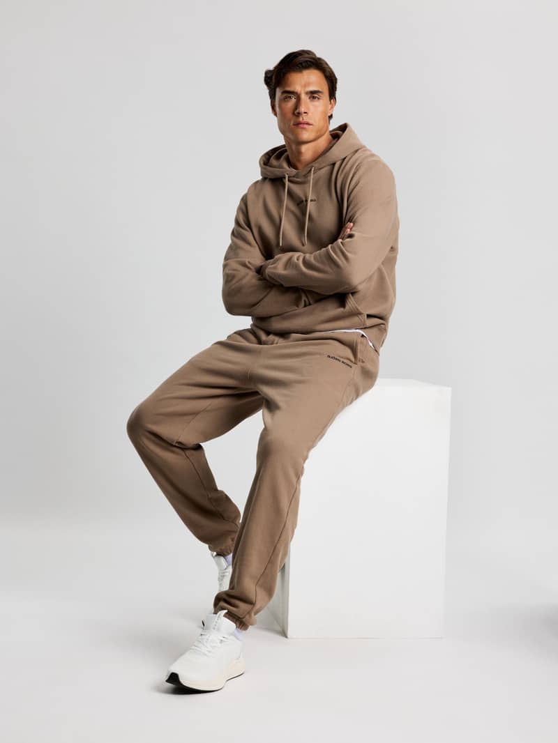 Man in a matching brown hoodie and sweatpants sits on a white cube, arms crossed, against a plain white background.