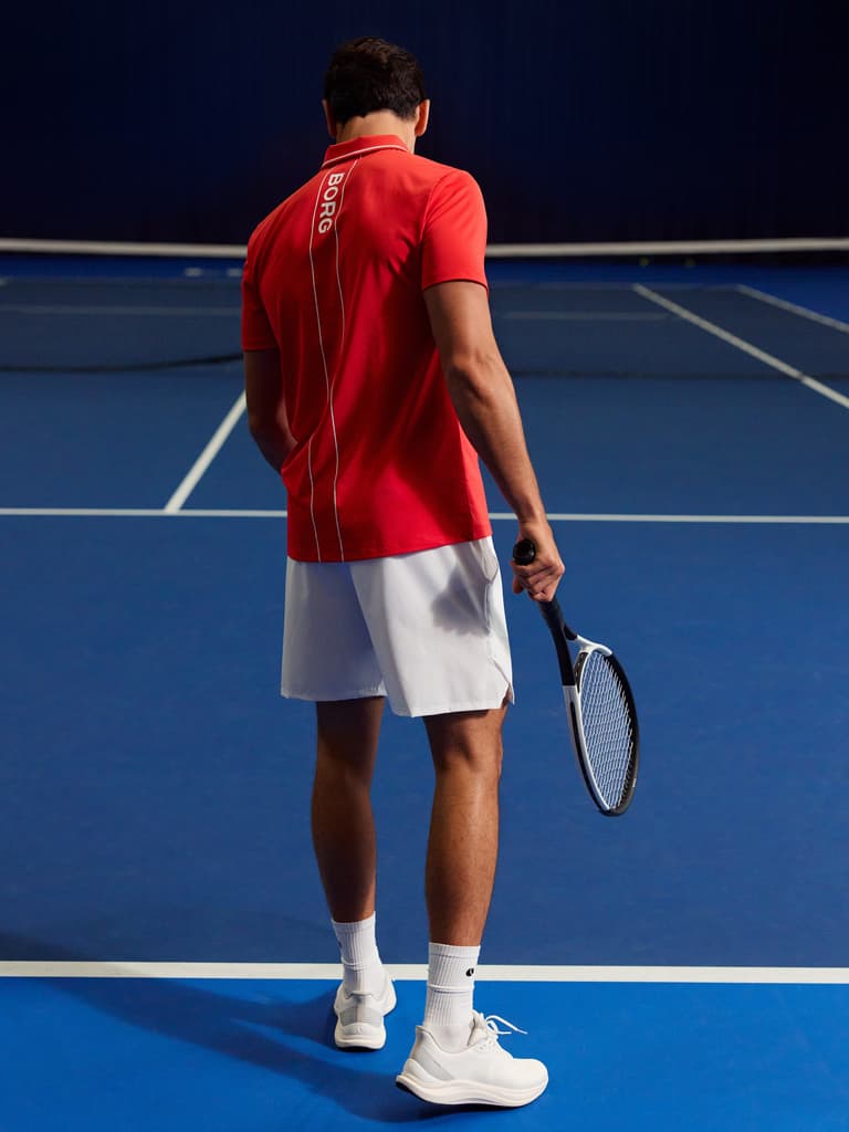 Man in red shirt and white shorts holding a tennis racket, standing on a blue tennis court with "We Know Tennis" text at the bottom.