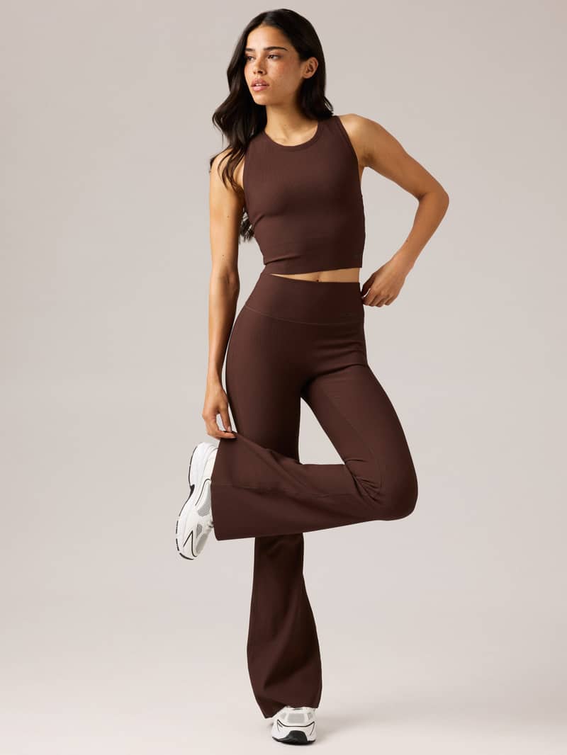 A person in a brown athletic outfit poses confidently with one leg slightly raised, gazing to the side, against a neutral background.