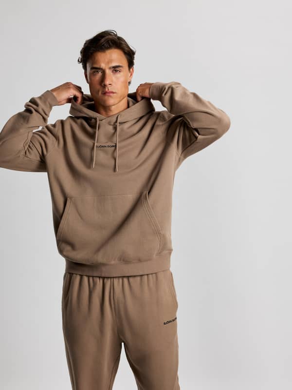 A person wearing a matching beige hooded sweatshirt and sweatpants adjusts the hood against a plain background.