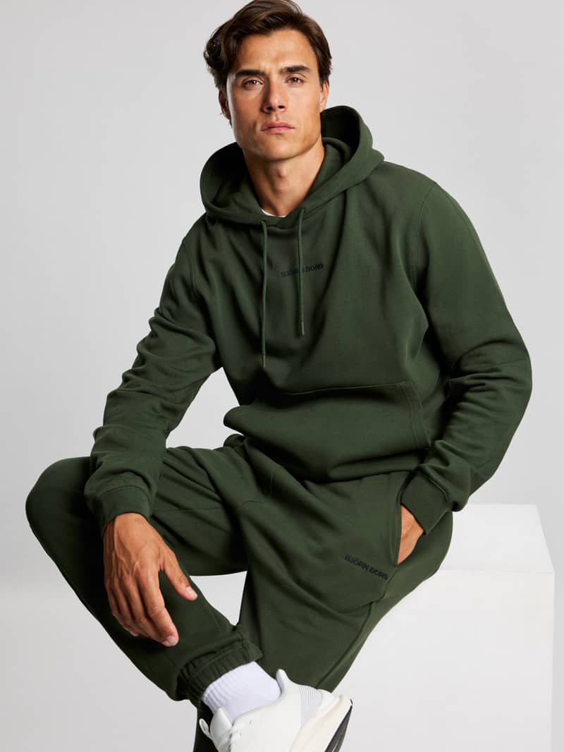 Man in a green hoodie and matching sweatpants seated, wearing white sneakers, looking confidently at the camera against a plain background.