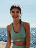 Smiling woman in a green sports bra and leggings stands by the sea on a sunny day, with sparkling water in the background.