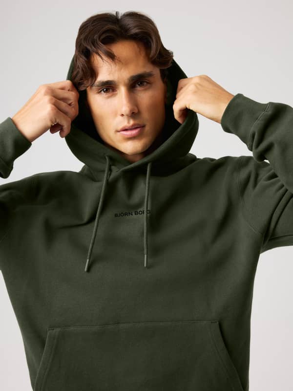 A person in a green hoodie adjusts the hood while looking at the camera against a plain background. The hoodie has "BJÖRN BORG" text on the front.