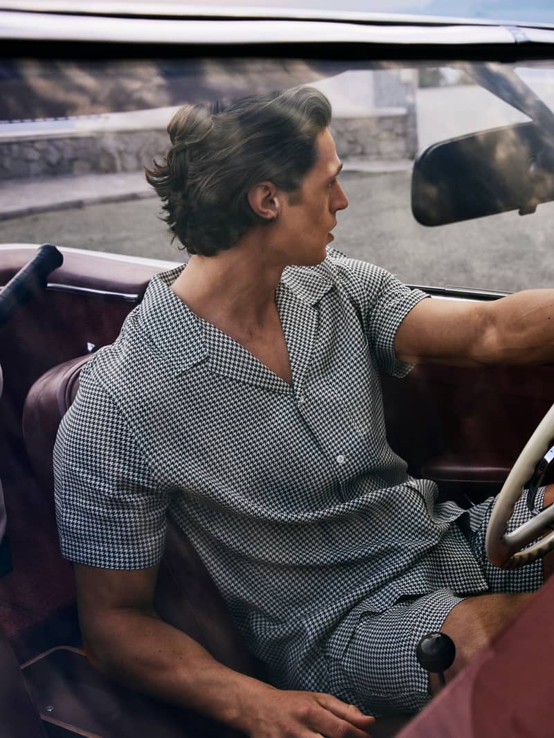 Man in a checkered outfit driving a classic car, gazing out the window on a sunny day.