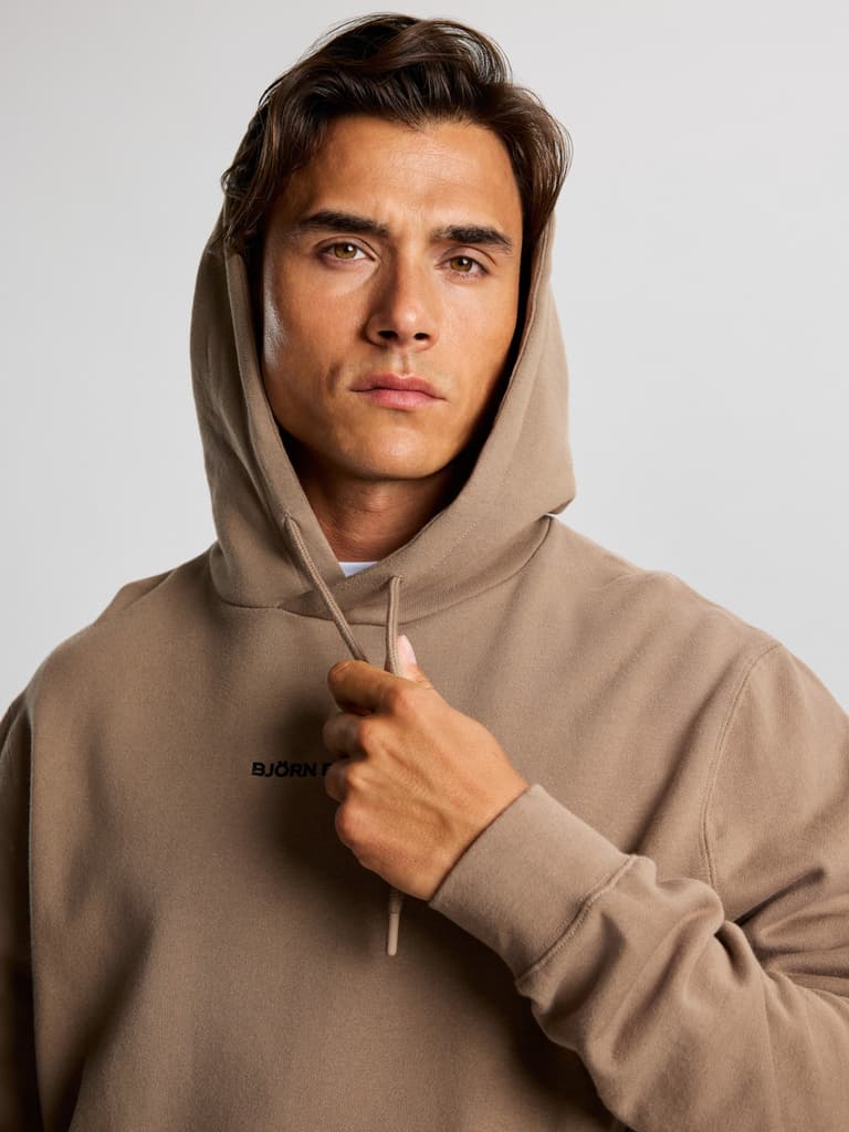 Person wearing a beige hoodie, pulling the hood with one hand, looking directly at the camera against a plain background.